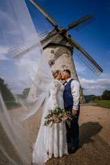 A bride and groom share a romantic kiss in front of an old windmill on a sunny day. Her veil flows in the wind and she holds a bouquet of pastel flowers.
