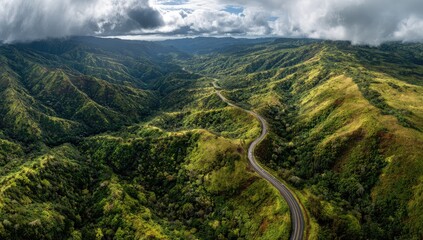 Aerial view of a winding road snaking through a lush, verdant valley nestled within a range of rolling green mountains under a dramatic, partly cloudy sky