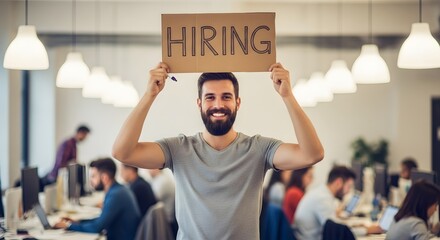 A smiling man with a beard holds a "HIRING" sign in a modern, open-plan office filled with working professionals.