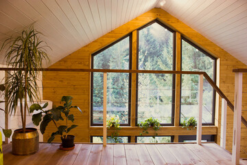 A cozy wooden cabin loft with a large window overlooking a lush green forest. Interior view of a...