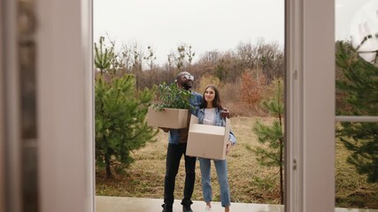Happy young couple carrying cardboard boxes outdoors near new house, smiling man and woman moving together, relocation, real estate purchase, eco living and new home ownership lifestyle concept