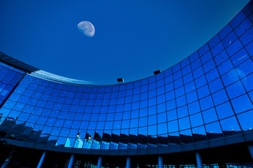 Curved glass facade of modern office building reflecting moon in vibrant blue night sky