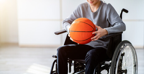 Cropped view of disabled teenager in wheelchair holding basketball at home. Unrecognizable...