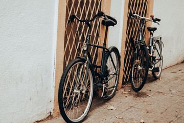 Two vintage bicycles lean against a textured wall and ornate metal grate. The bikes have a classic...