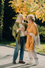 Two senior women smiling and holding coffee cups in autumn park