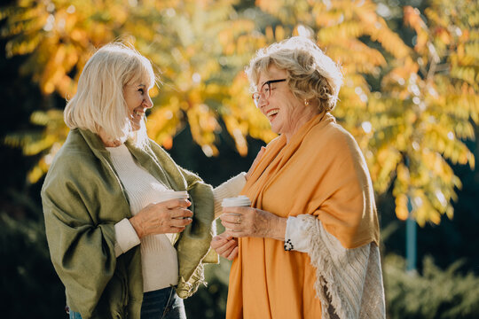 Two senior women laughing together while drinking coffee in autumn park