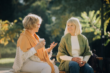 Two senior women talking and drinking coffee on a bench in a park