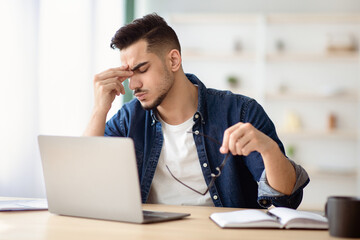 Exhausted arab guy sitting at workdesk with laptop in office, removing glasses and rubbing nose...