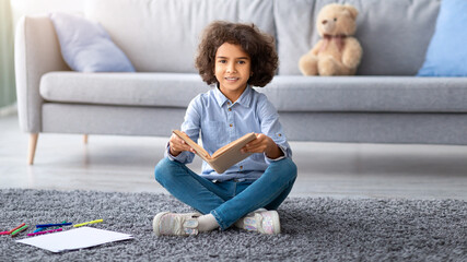Enjoying Interesting Story. Portrait of cheerful smiling black girl holding and reading paper book, sitting on the floor carpet in living room, copy space. Teenager looking at camera, modern interior