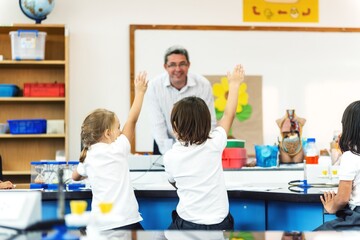 Children in a classroom raising hands, engaging with a teacher. Classroom interaction with elementary students and teacher. Learning environment with active participation. Diverse elementary students.