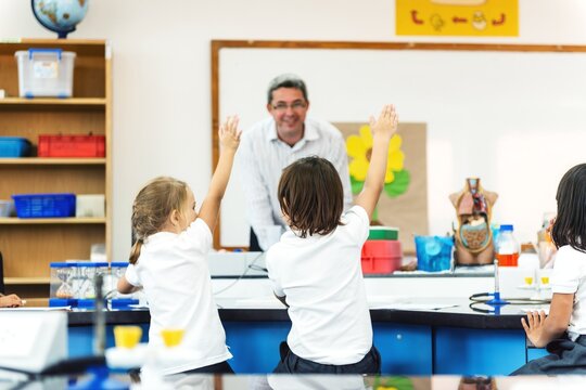 Children in a classroom raising hands, engaging with a teacher. Classroom interaction with elementary students and teacher. Learning environment with active participation. Diverse elementary students.