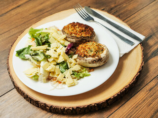 A plate of food with a fork and knife on a wooden table. The plate has a fresh salad with cheese and two fancy posh stuffed mushroom patties. Simple tasty meal.