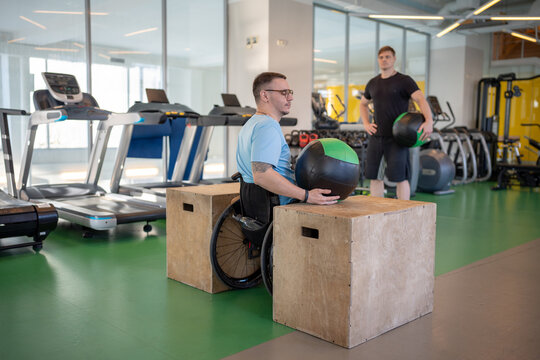 Determined man in wheelchair do guided therapy sports, lifting and balancing heavy medical ball under personal trainer supervision. Male focused on shoulder stability core activation, spinal trauma - Powered by Adobe