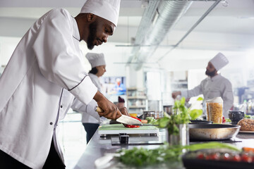 African american male chef slicing a red bell pepper on a cutting board with stainless steel tools in a restaurant kitchen. Man chopping delicious ingredients for a new gourmet dish.