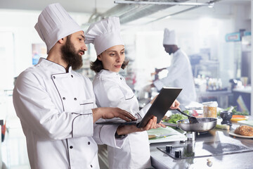 Team of two chefs researching recipes for new gourmet dishes on laptop, preparing meals in modern equipped restaurant kitchen. Professionals work on food prep, fresh produce ingredients.