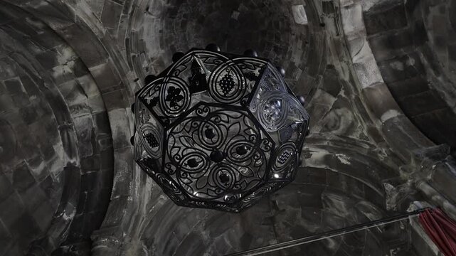 A detailed, close-up view of an antique black wrought iron chandelier with multiple bulbs, showcasing its intricate scrollwork and decorative design against a dimly lit, textured background.