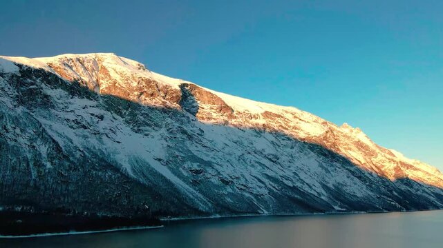 This drone footage captures the serene beauty of the Eresfjord and Eikesdalen region in Norway during golden hour, showcasing snow capped mountains bathed in warm sunlight reflecting on the calm fjord