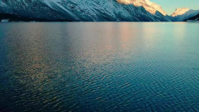 This drone footage captures the serene fjord waters reflecting the majestic, snow capped mountains bathed in the warm glow of sunrise. Ideal for showcasing natural beauty, travel, tranquility, and Nor