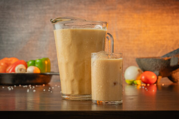 Glass and jar of traditional cold iced barley water on wooden table with kitchen equipment and ingredients in the background