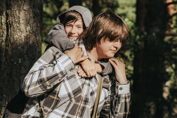 A cheerful boy in a hoodie rides on the back of an older boy in a plaid shirt, sharing a playful piggyback moment outdoors in the sunlight.
