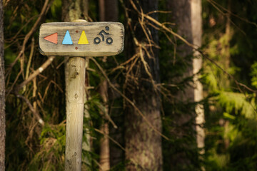 A wooden trail sign with colorful triangle symbols and a cyclist icon, marking a biking path through the forest.