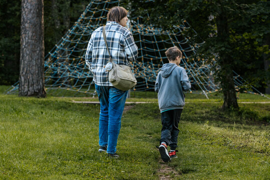 A man and a boy walk together on a grassy path in a park, passing by a large rope climbing structure among the trees.