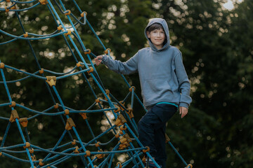 A boy in a grey hoodie carefully balances on a rope climbing structure, arms stretched wide for stability.