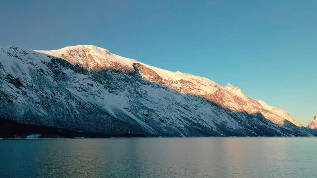 This drone footage captures the serene beauty of a snow covered mountain range reflecting in calm fjord waters during a golden sunrise.