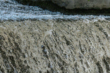 Cascading water flows over a concrete ledge, creating textured ripples and foamy currents beneath...