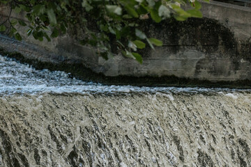 Cascading water flows over a concrete ledge, creating textured ripples and foamy currents beneath...