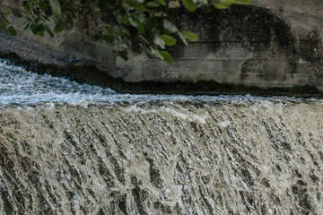 Cascading water flows over a concrete ledge, creating textured ripples and foamy currents beneath leafy branches.