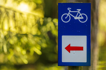 A blue bicycle path sign with a red arrow pointing left, mounted on a wooden post in a forest,...