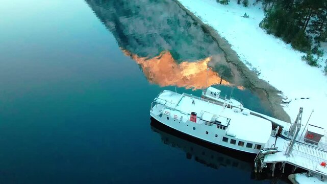 Aerial footage captures the serene beauty of Eresfjord or Eikesdalen in winter. A tranquil boat rests by the snow covered shoreline, reflecting the warm hues of the setting or rising sun on the water'