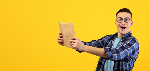 Surprised male student holding textbook at arm distance, looking at camera in shock on orange...