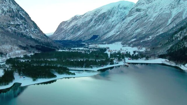Aerial drone footage capturing the serene beauty of a snow covered Norwegian valley, featuring a calm lake surrounded by majestic mountains and evergreen forests. This peaceful winter landscape could 