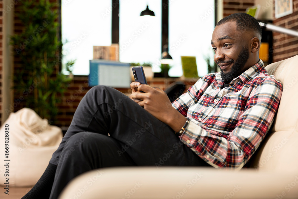 Poster black businessman resting on couch, reading text messages on smartphone and catching up with family 