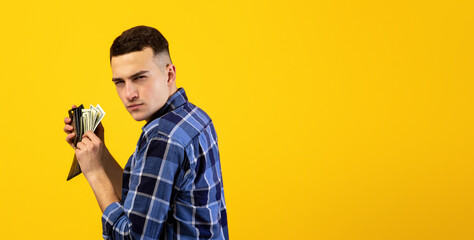 Young man with wallet full of money being greedy over his wealth on orange studio background. Millennial guy with dollars acting mean and stingy, not willing to pay taxes or share his income © Prostock-studio