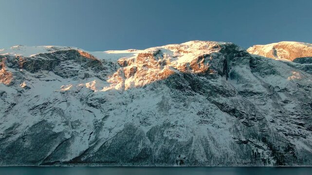 aerial footage captures the majestic snow covered mountains of Eresfjord or Eikesdalen in Norway, bathed in the warm, ethereal light of the golden hour.