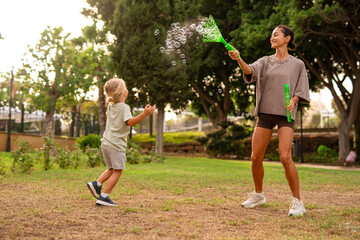 Happy mother playing with her young son in the park, creating soap bubbles with a wand, both...
