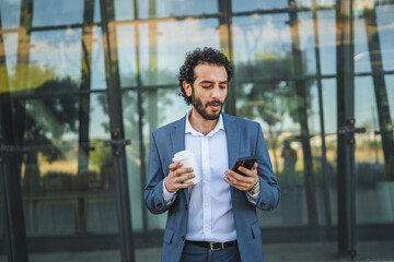 Businessman drinking coffee and reading news on smart phone in front of office building