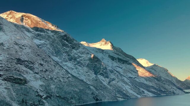 Aerial footage captures the beauty of a snow covered mountain range meeting a serene fjord under a clear blue sky, with golden hour sunlight illuminating the peaks. Ideal for travel, nature, documenta
