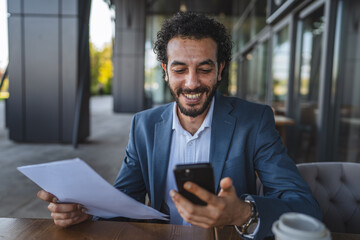 Happy businessman reading documents and using smart phone in outdoor cafe