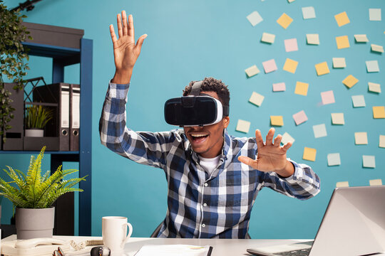 Excited employee uses VR headset, blending technology with workplace creativity. African american man exploring virtual reality, fully engaged in an interactive digital world at his office desk.