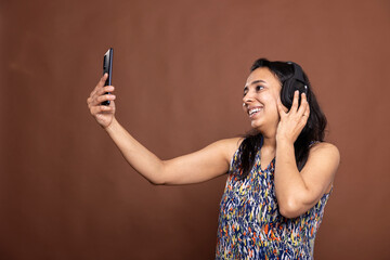 Young indian woman with bright smile listens to music while holding cellphone for a selfie vlog....