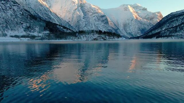 aerial or ground footage capturing the serene beauty of Eresfjord or Eikesdalen in winter. Snow-covered peaks rise majestically, bathed in golden hour light, with their stunning reflections shimmering