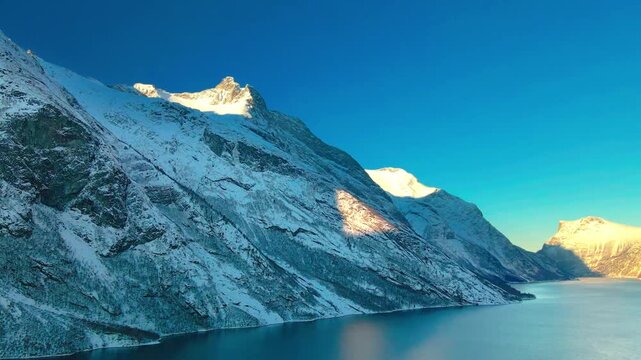 aerial or wide shot footage capturing the serene beauty of a snow covered fjord landscape, potentially Eresfjord or Eikesdalen in Norway, bathed in the warm glow of sunrise or sunset.