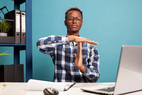 Male business consultant making a timeout symbol with his arms, signaling a break from work responsibilities. African american manager seated at office desk, using his hands to gesture a pause sign.