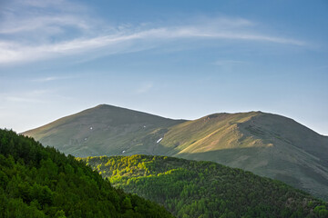 Fototapeta premium Peaceful mountain landscape with lush green forests at the base and sunlit slopes under a clear sky. The scene conveys natural beauty, serenity and the majesty of untouched wilderness.
