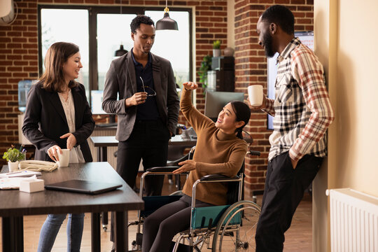 Professional employees having a lighthearted conversation, promoting teamwork in a business setting. Cheerful asian woman in a wheelchair chatting with her inclusive team, taking a short break.