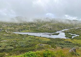 Moody mountain landscape outside Eidfjord, Norway, with fog drifting across rocky green highlands and a winding river cutting through the valley. Atmospheric Nordic wilderness scenery in summer.